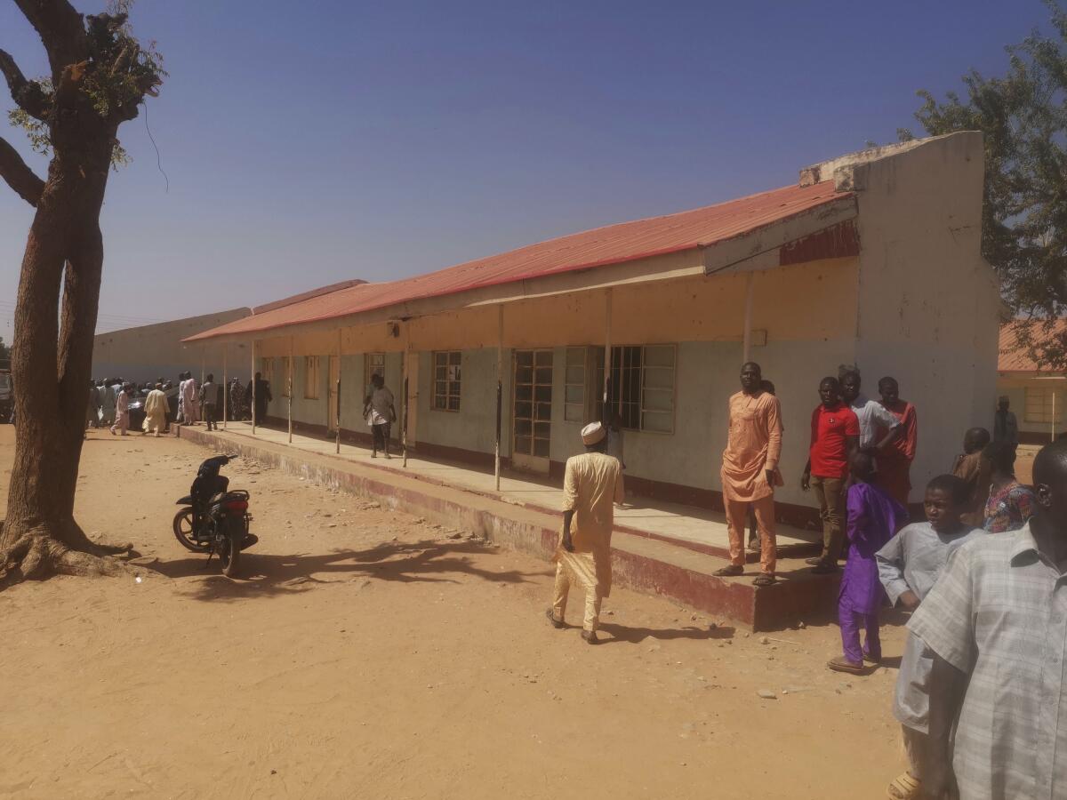 In a bare dirt courtyard, people gather outside a long low building. 
