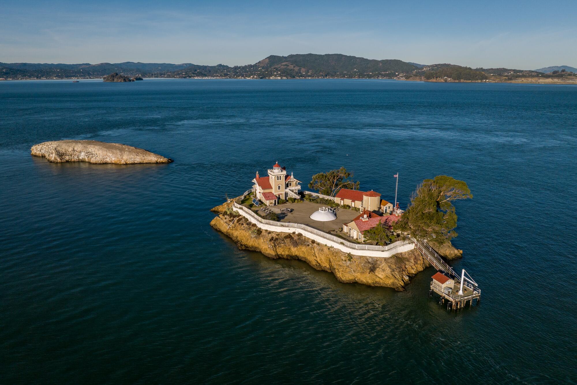 An island with a lighthouse and other buildings, with hills across the water in the background