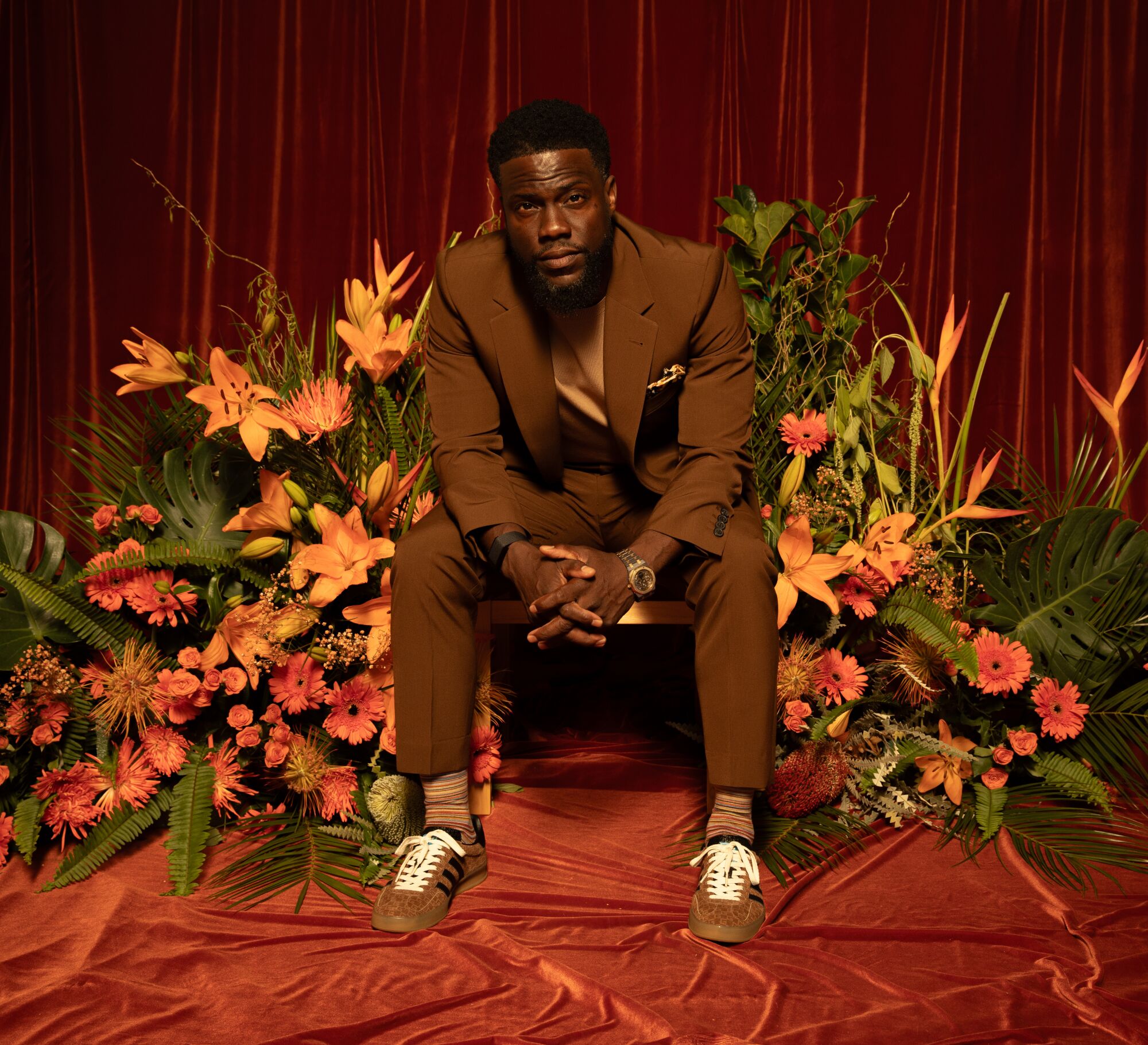Kevin Hart in a brown suit and shoes sitting among orange flowers and green leaves and a copper carpet
