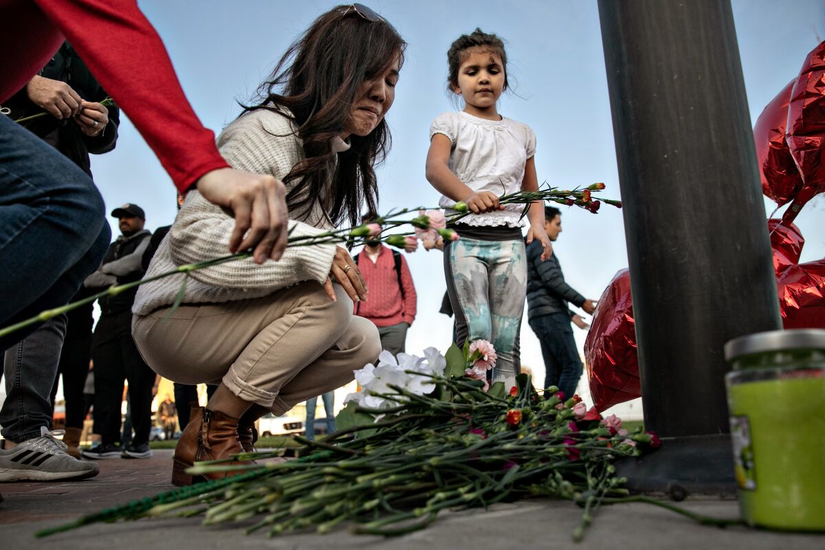 A woman in a red sweater cries as she crouches by a pile of flowers. A little girl holds more flowers.