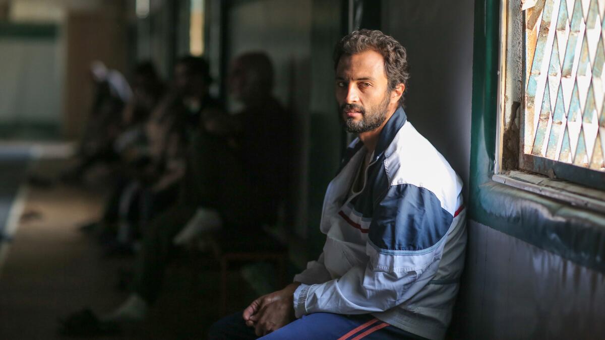 A bearded man sits on a bench in front of a window.