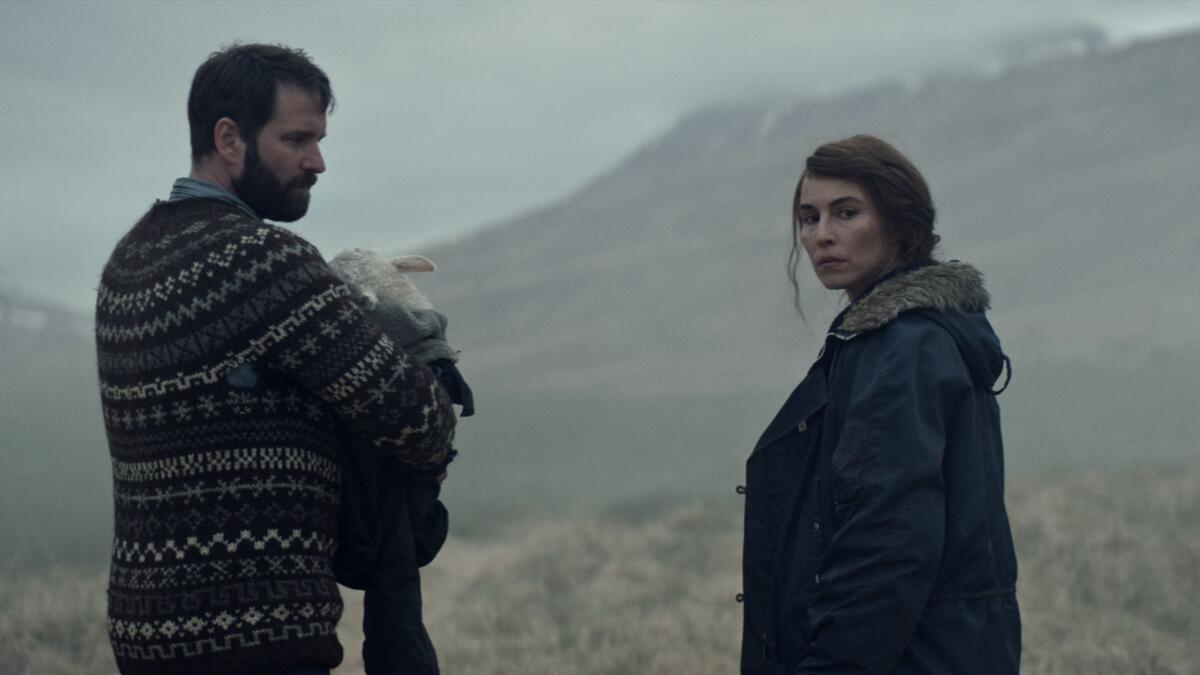 A man holding an animal stands with a woman in a field in the movie “Lamb.”