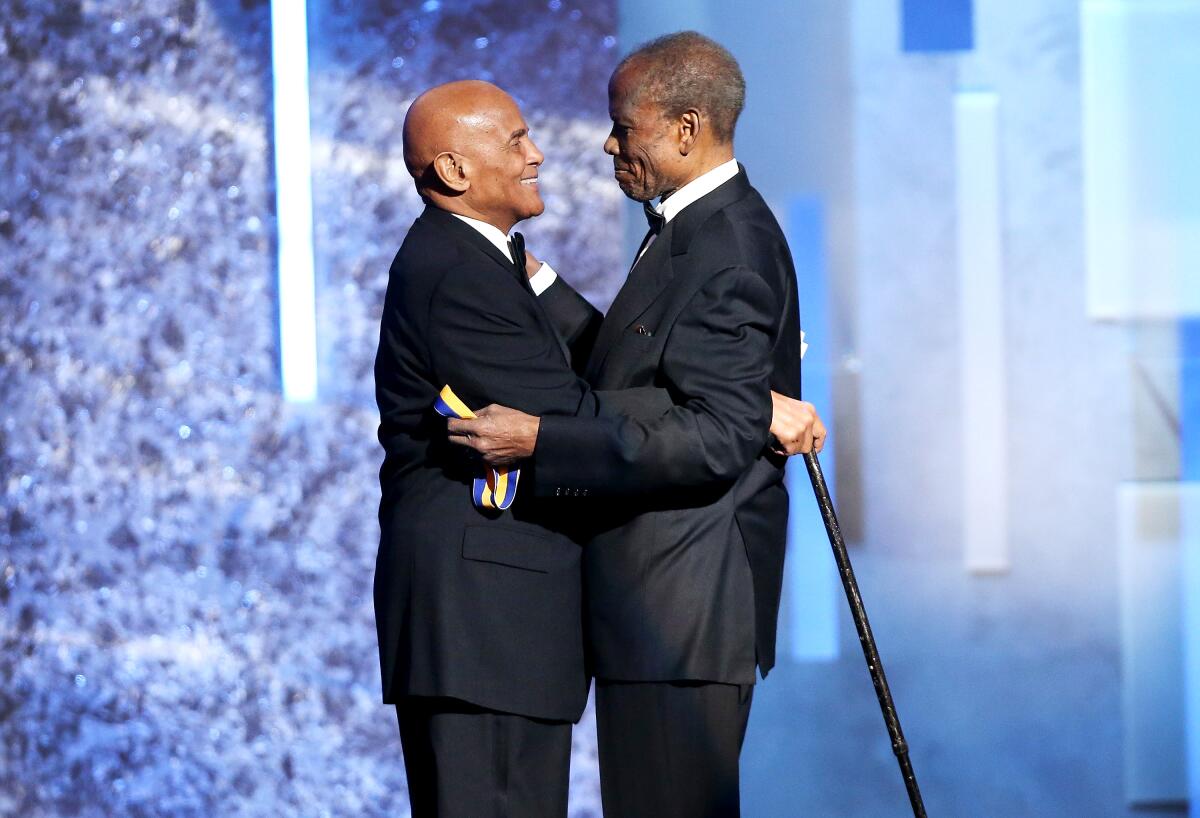 Sidney Poitier holds a medal and Harry Belafonte holds a cane as they embrace onstage.