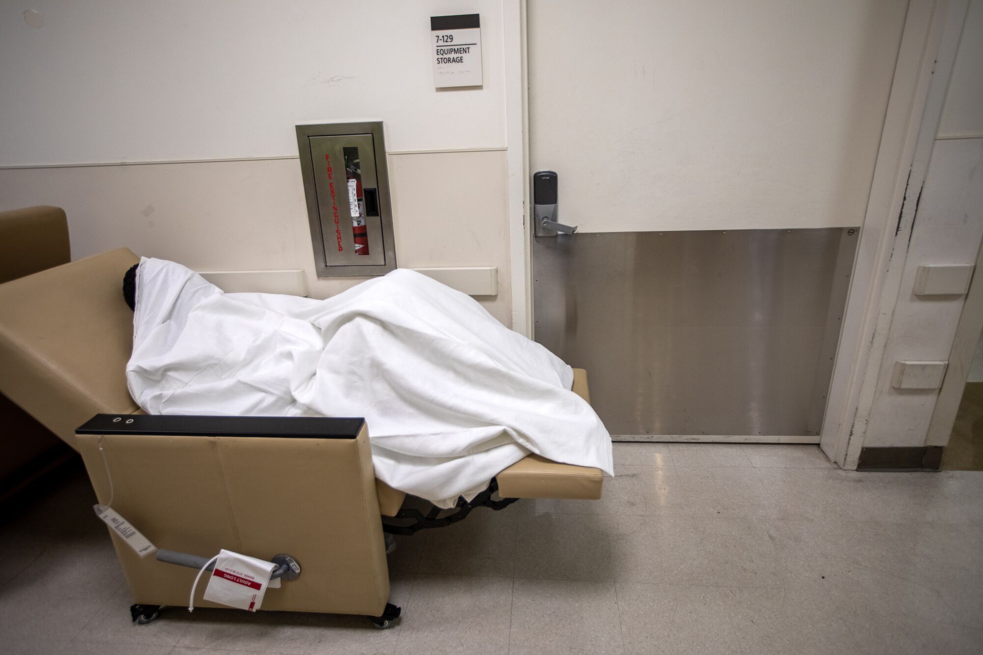 A hospital patient, covered with a blanket, rests on a chair in the hallway