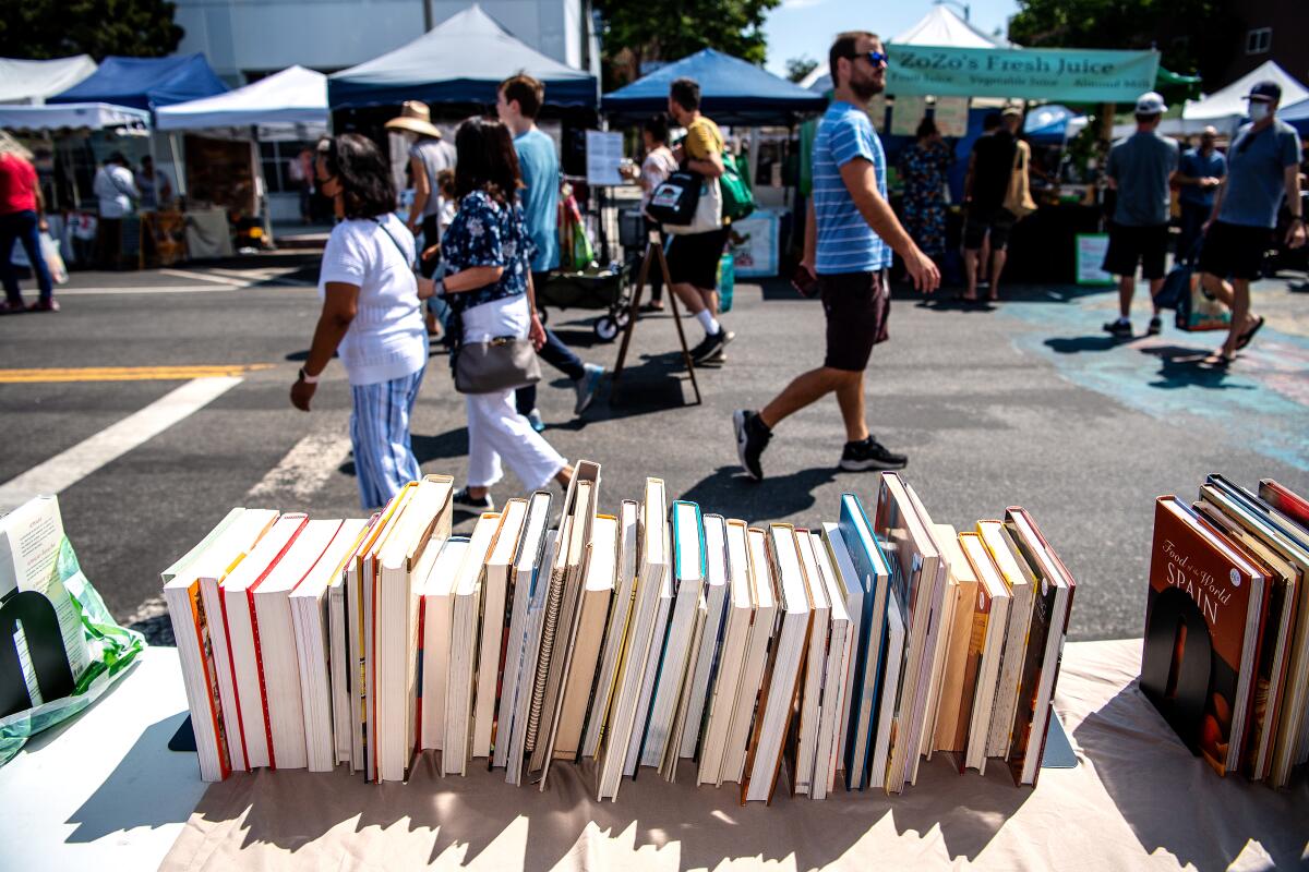 People walk past an array of cookbooks for sale on a table at an outdoor market.