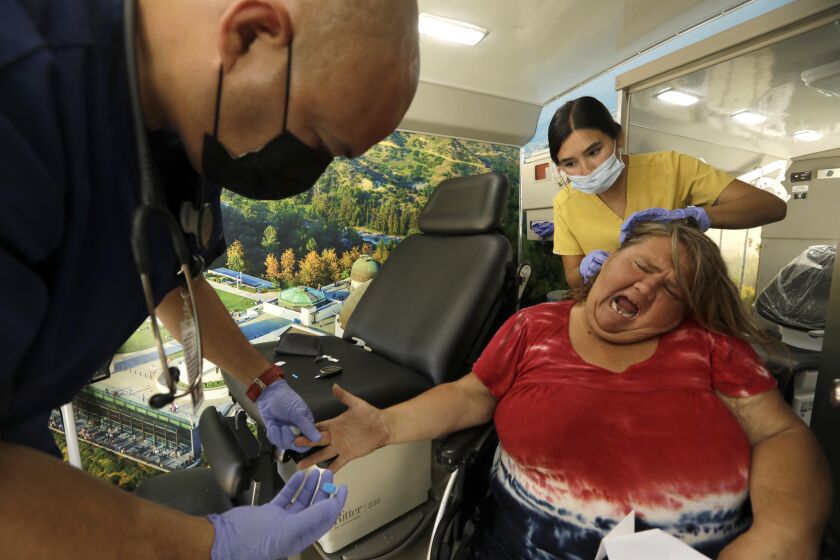 HOLLYWOOD, CA - JUNE 20, 2022 - - Homeless patient Lisa Rogers grimaces after a prick by Saban Community Clinic medical assistant Jeffrey Figueroa, left, who checks her blood sugar level, as medical assistant Samantha Kumpf looks for hair lice inside the Saban Community Clinic mobile unit parked in front of A Bridge Home shelter on Schrader Boulevard in Hollywood on June 20, 2022. The new Saban Community Clinic mobile unit visits and treats homeless patients who are currently living in transitional housing and treat their medical conditions. (Genaro Molina / Los Angeles Times)