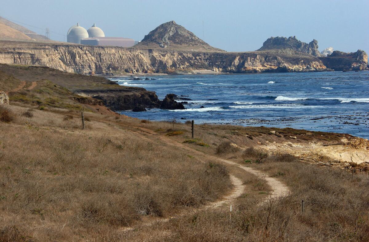 A view of a grassy dirt road, ocean, cliffs and, in the distance, reactor containment structures.