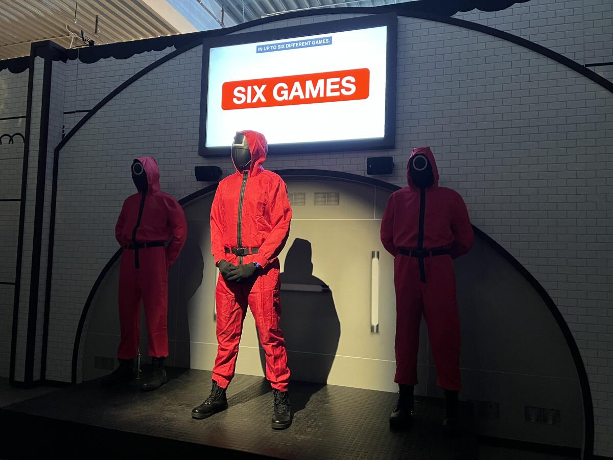 Three red-suited background actors stand in front of the orientation room at Squid Game: The Trials.