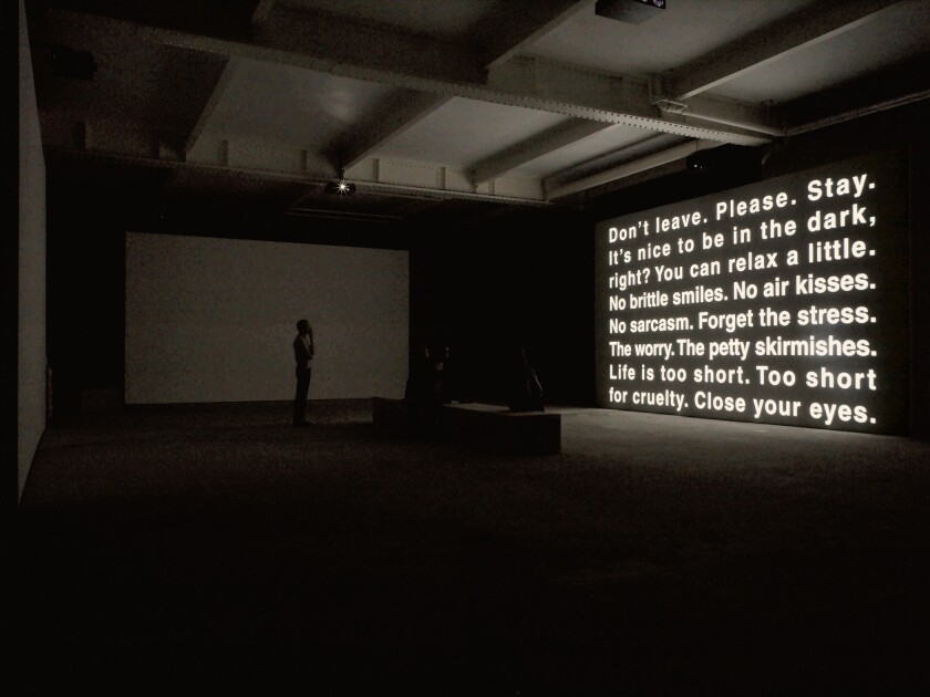 A person stands in a darkened gallery looking at text on a video screen that begins: "Don't leave. Please. Stay."