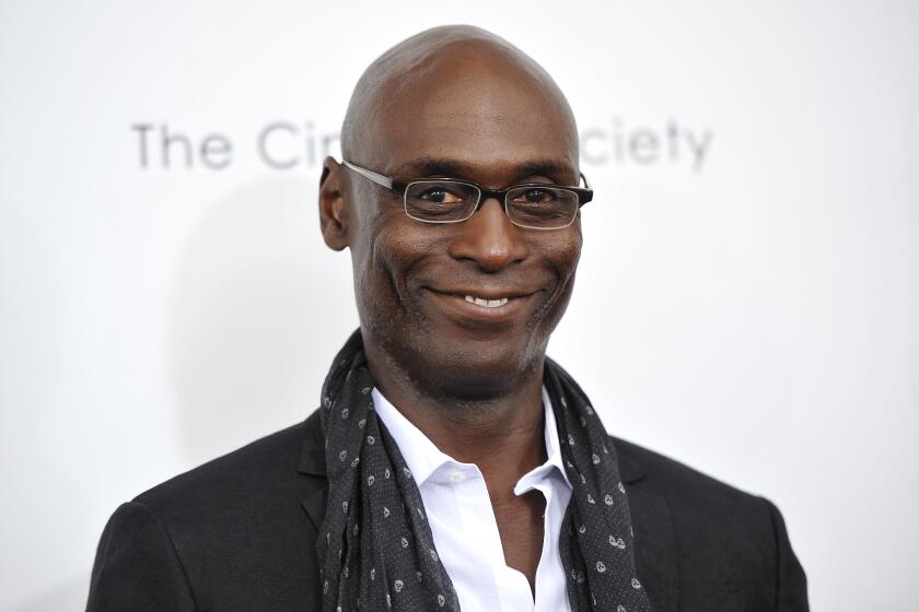 Lance Reddick smiles in a black suit, scarf and glasses