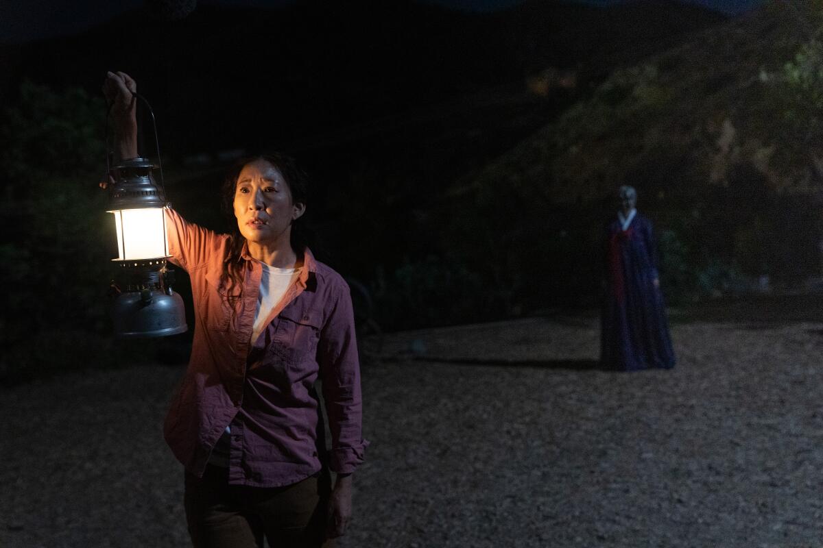 A woman, left, holds up a lantern in the dark with an eerie figure of the woman to the back right 
