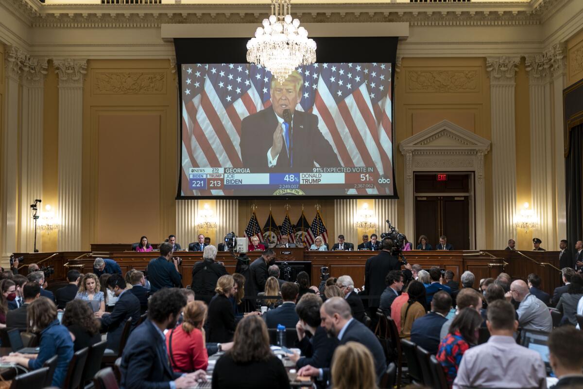 A video of former President Trump plays on a large screen in a crowded hearing room