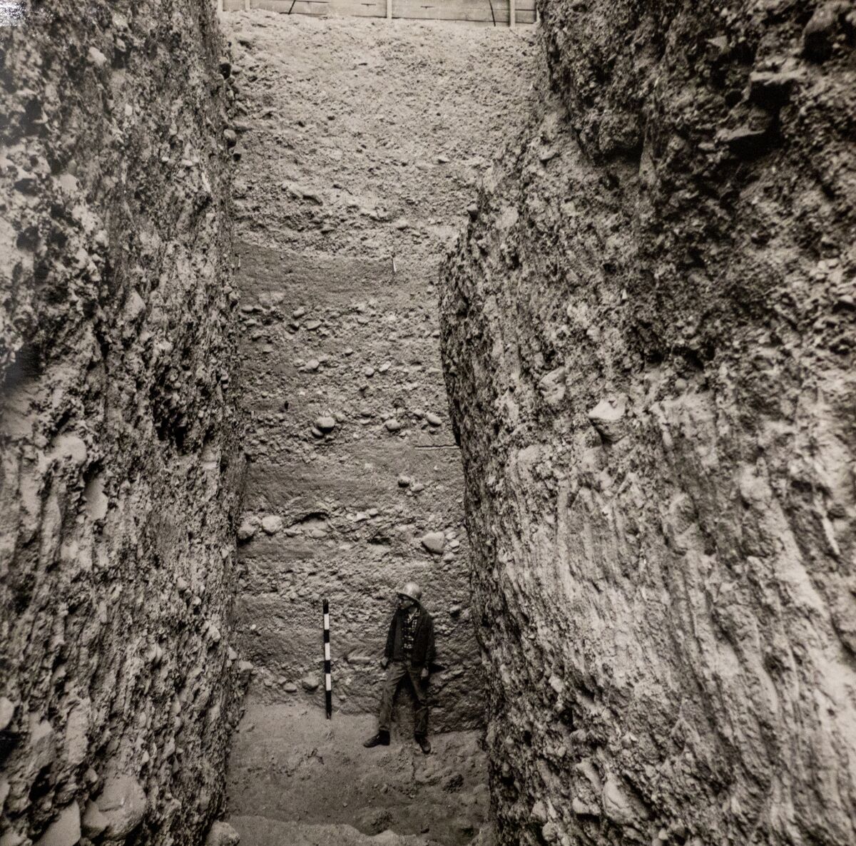 An old photo of a man standing in a deep, archaeological pit.