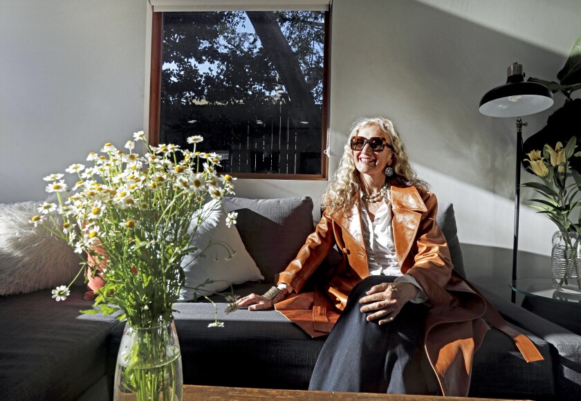 A woman sits on a couch with a tree visible through the window behind her.