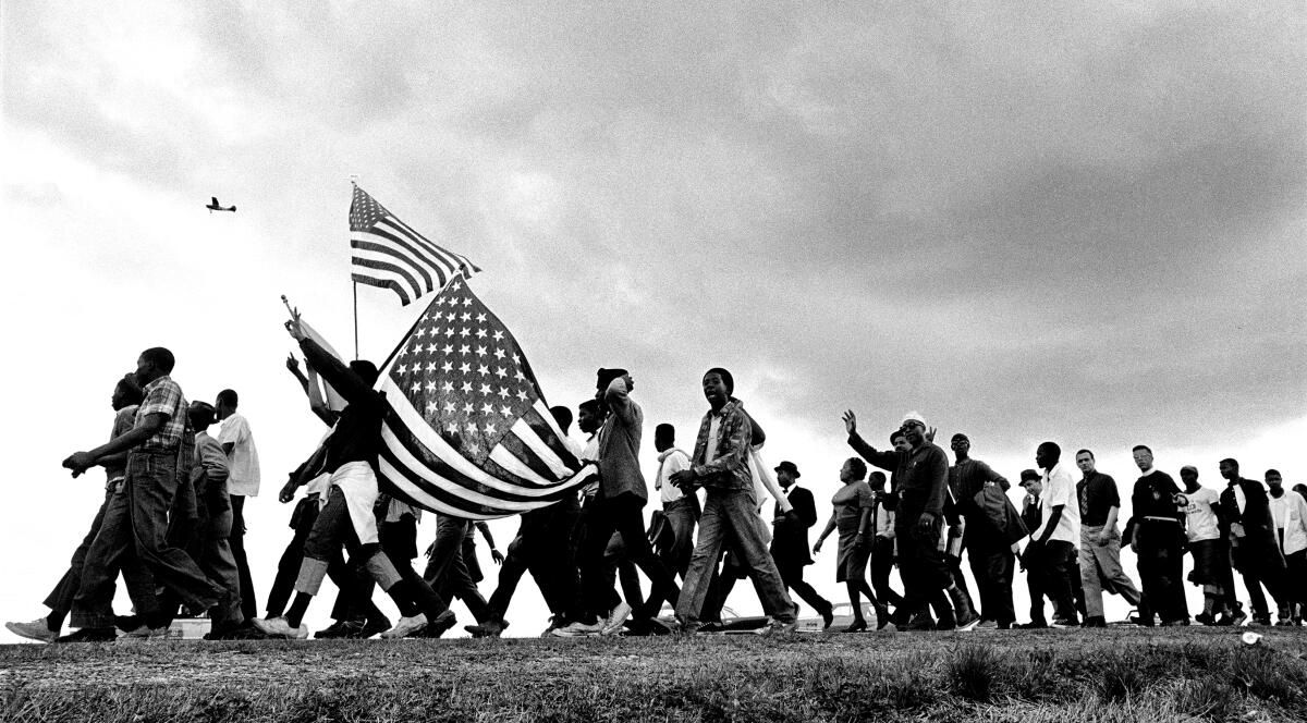 A black-and-white photo of people marching to the left, holding up U.S. flags.