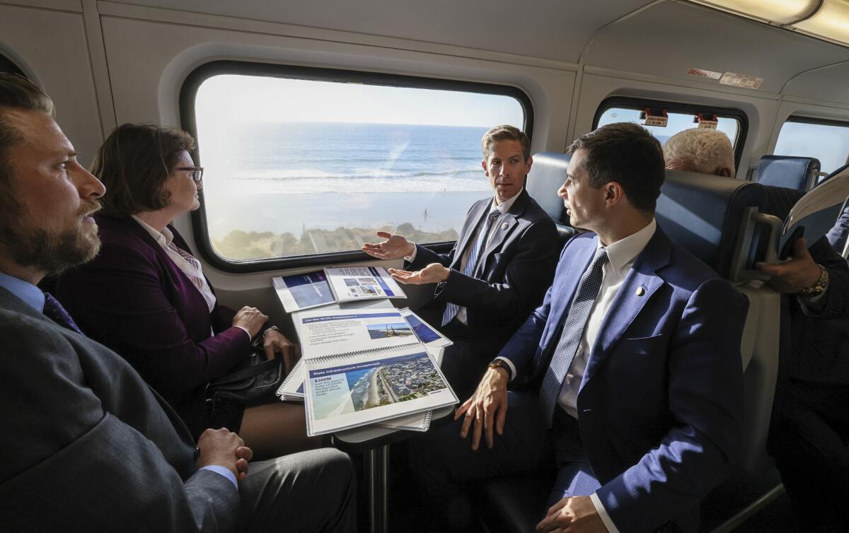 Three men and a woman, all wearing suits, sit facing each other on a passenger train with the ocean visible out the window.