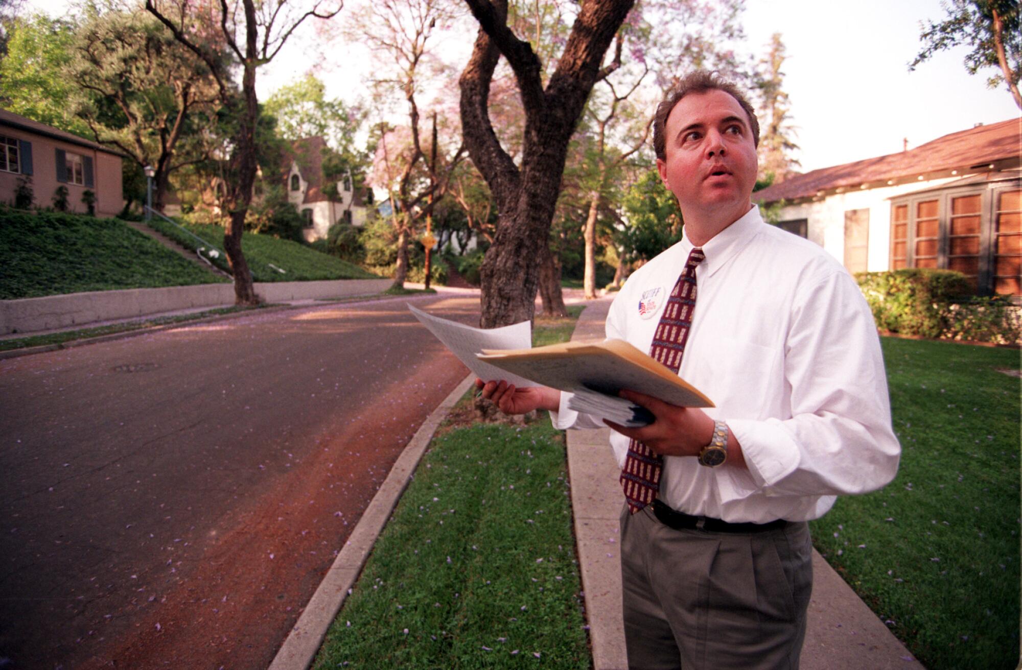 Adam Schiff holding a folder and papers and looking around as he stands on a sidewalk on a tree-lined residential street.