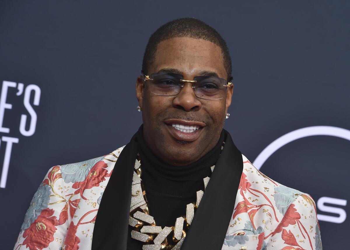 Busta Rhymes in a pink and blue patterned blazer, chain and sunglasses smiling in front of a dark backdrop