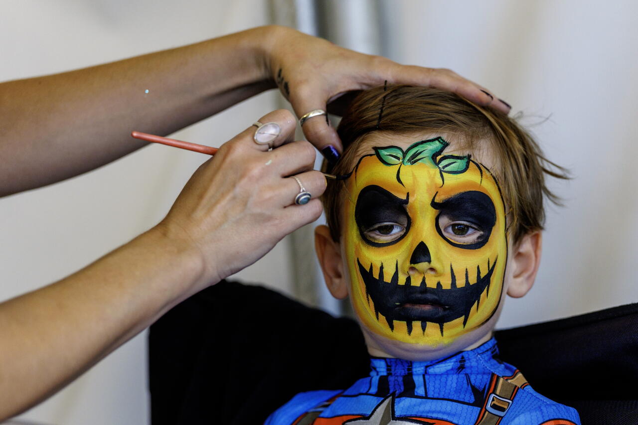 A hand paints a Jack-O-Lantern on a child’s face.