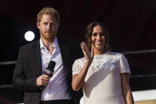 Prince Harry holds a microphone while wearing a suit and standing next to Meghan, who smiles and waves.