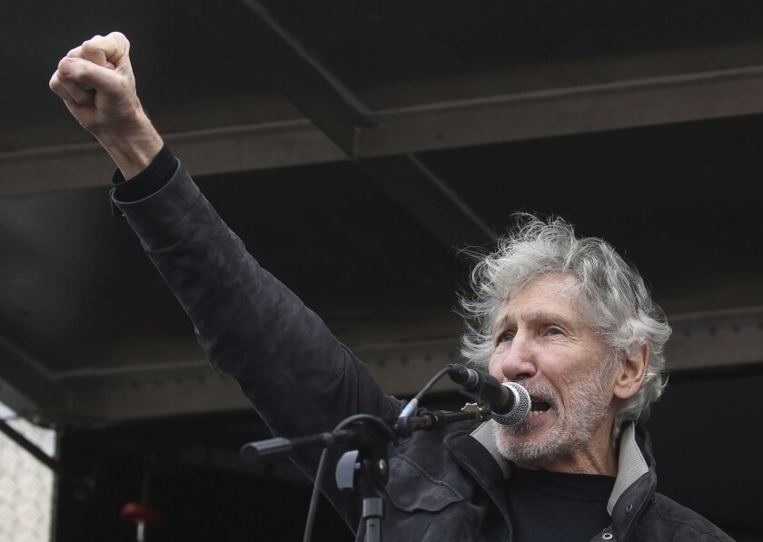 A man with gray hair and a scruffy beard speaks at a microphone while raising his right hand in a fist