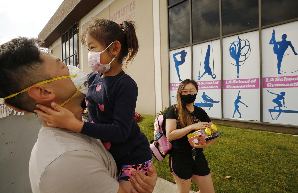 A father holds his young daughter while her mother carries supplies as they drop her off for day camp. All wear face masks.