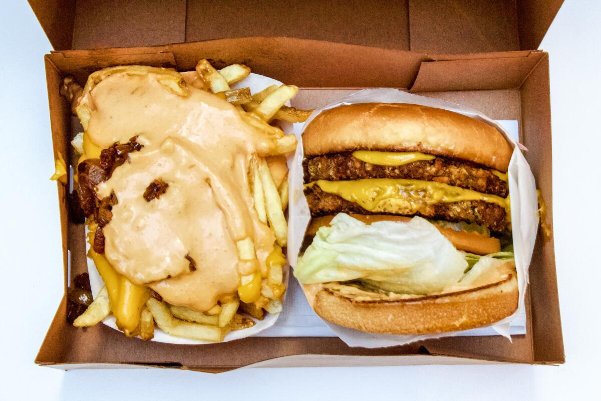 A vegan cheeseburger in a takeout cardboard tray next to sauce-covered french fries