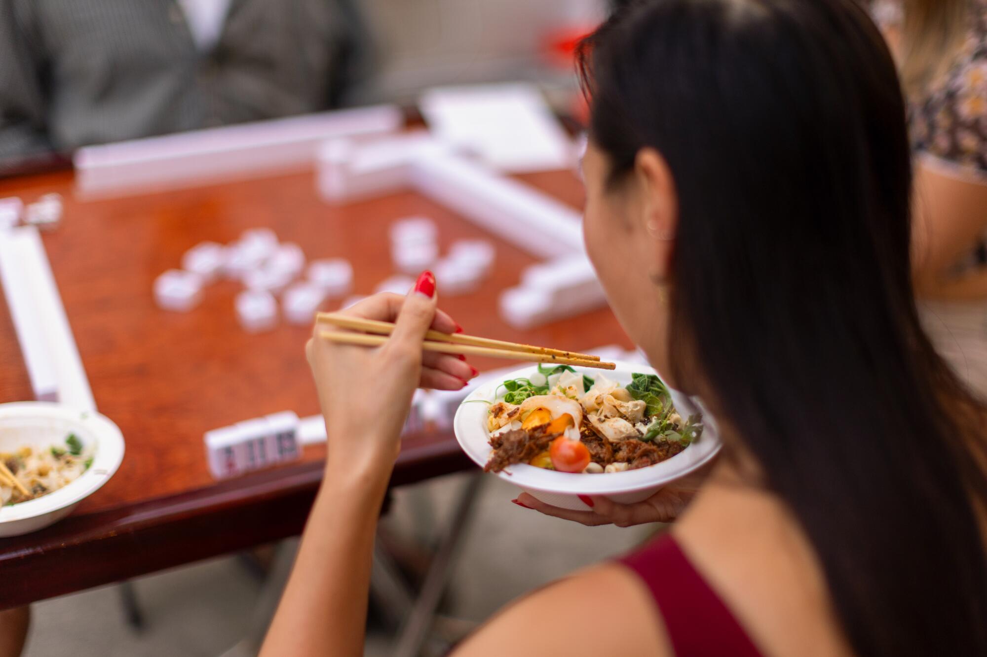 Una mujer come con palillos mientras está sentada en una mesa de mah-jongg.