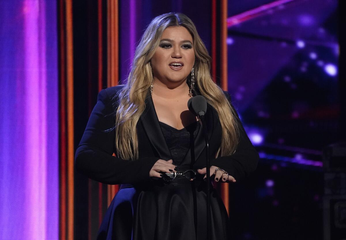 Kelly Clarkson with dark makeup in a black long-sleeve dress standing on a stage speaking into a microphone