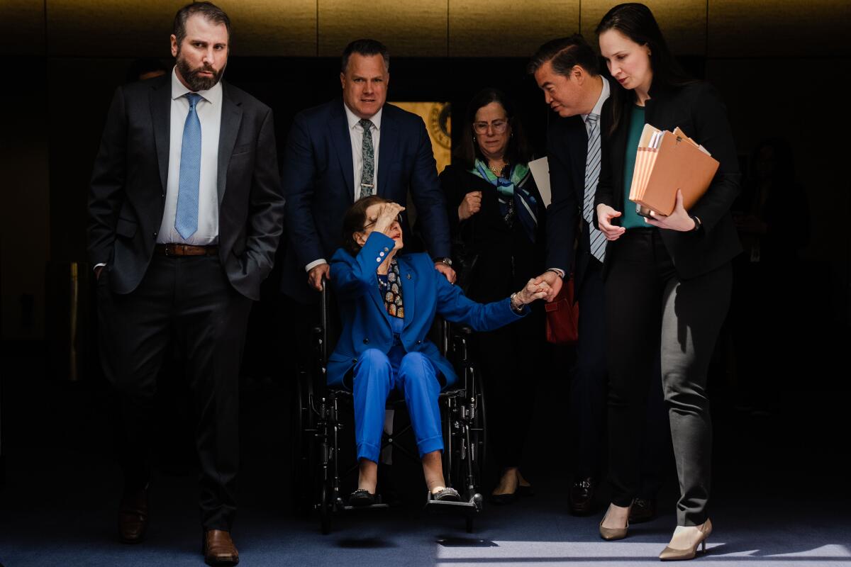 Dianne Feinstein, in a wheelchair flanked by staff, shields her eyes from the sun as a man takes her hand and talks with her.