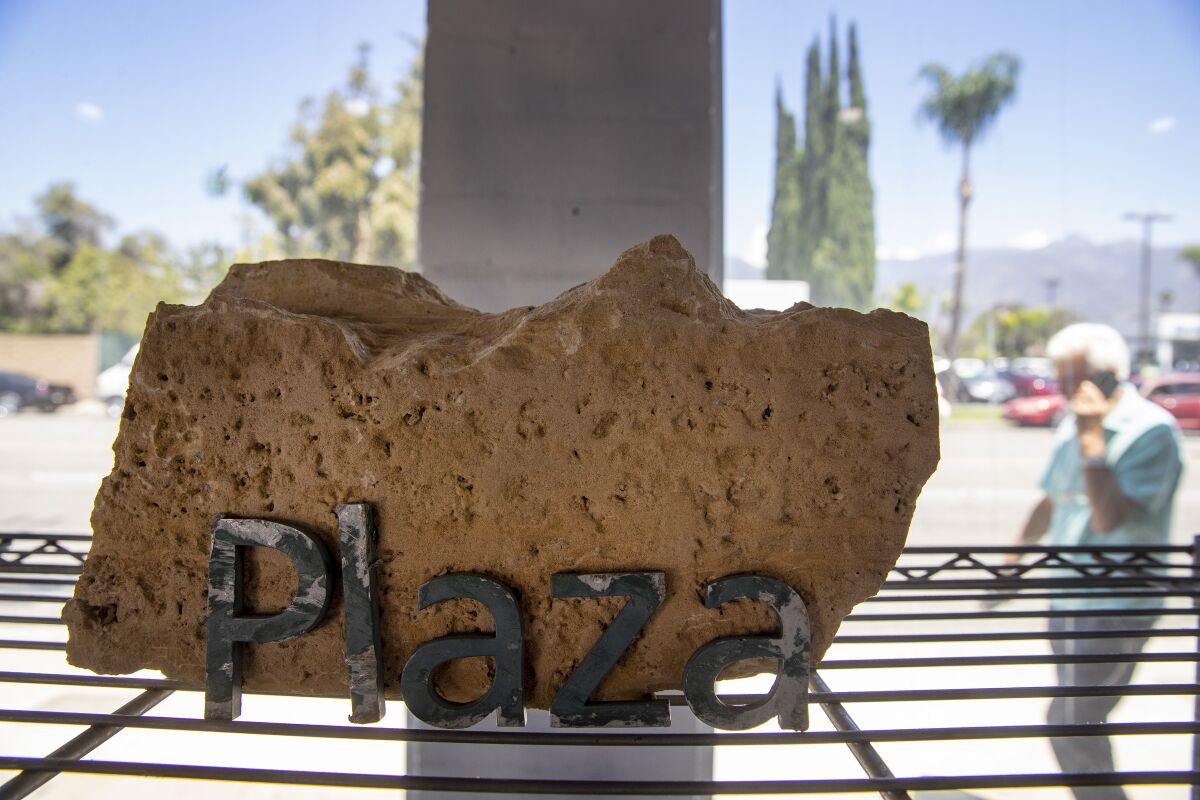 A fragment from LACMA's demolished buildings with a sign that reads "Plaza" sits on a metal shelf before a window