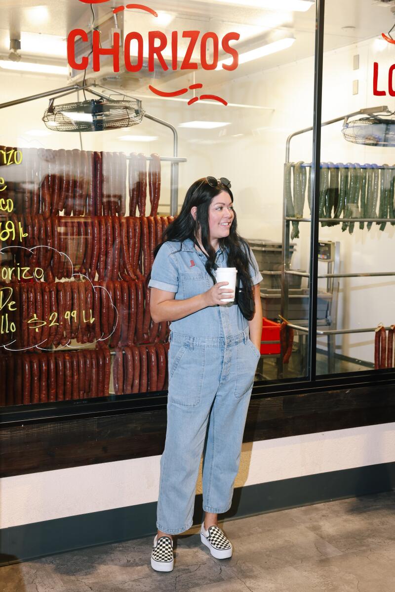 A woman in a denim jumpsuit stands in front of a window display of chorizo, holding a beverage.