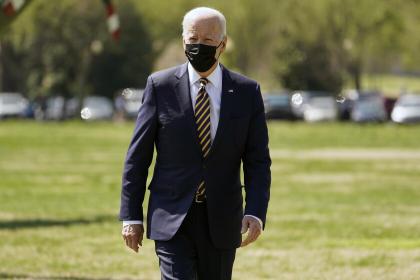 President Biden, wearing a face mask, walks on a lawn.