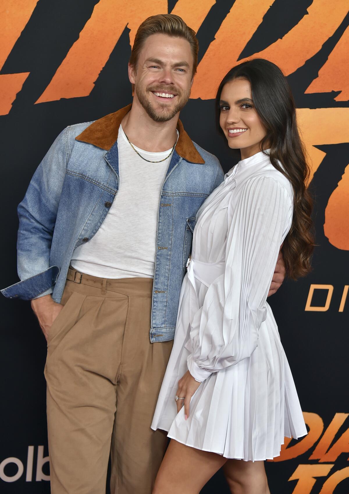 Derek Hough dressed in denim jacket poses next to Hayley Erbert in a white dress as the pair embrace and smile