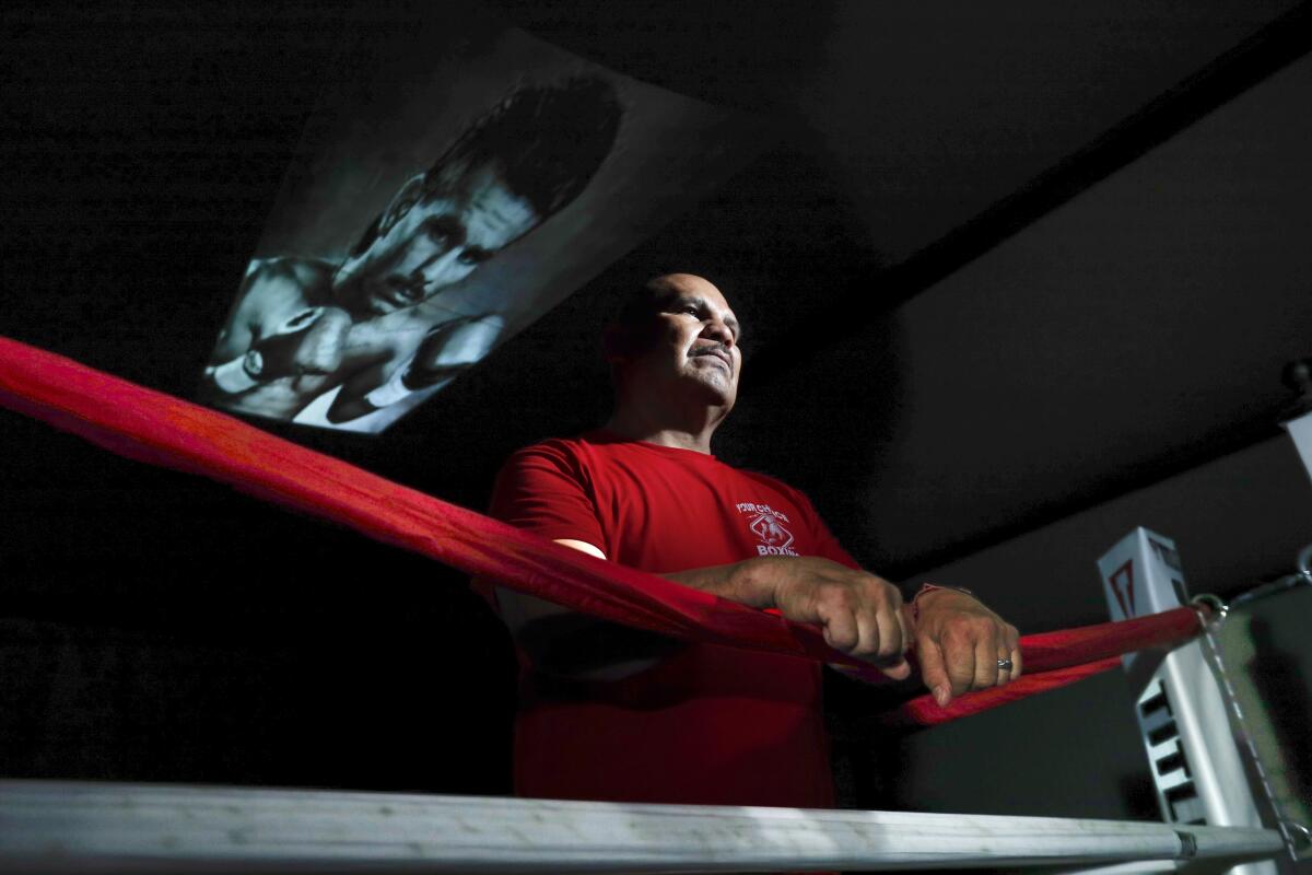 A man in a red shirt stands at the corner of boxing ring below a photo of his younger self
