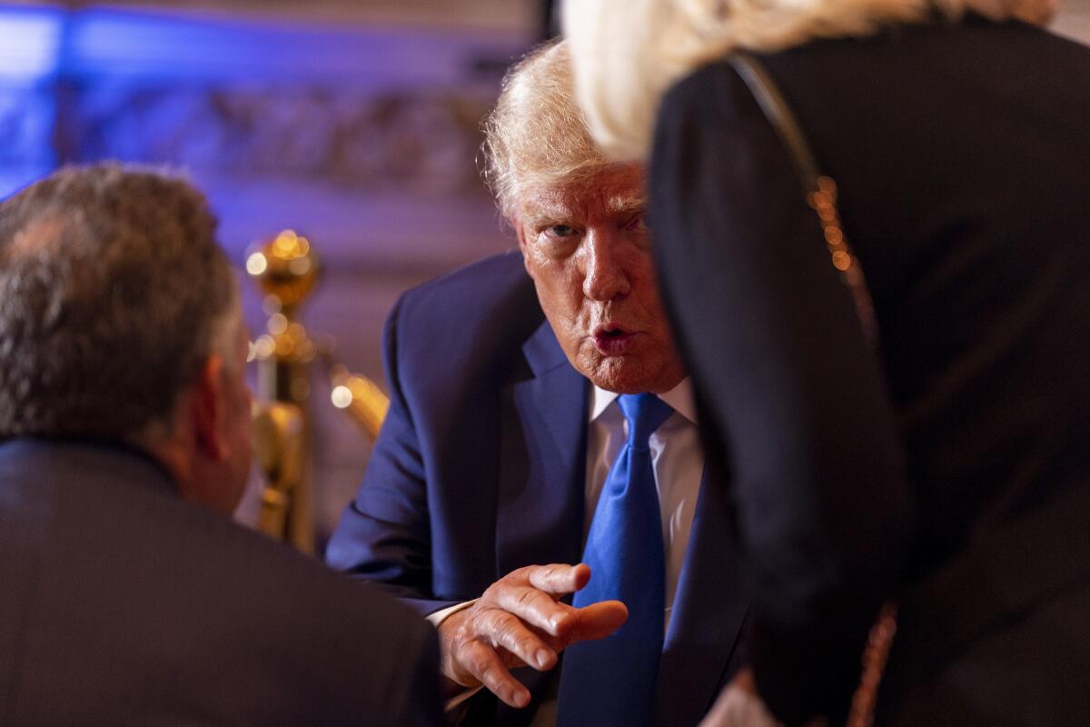 Donald Trump is seen leaning forward, gesturing with his right hand, as he talks to man seated across a table from him.