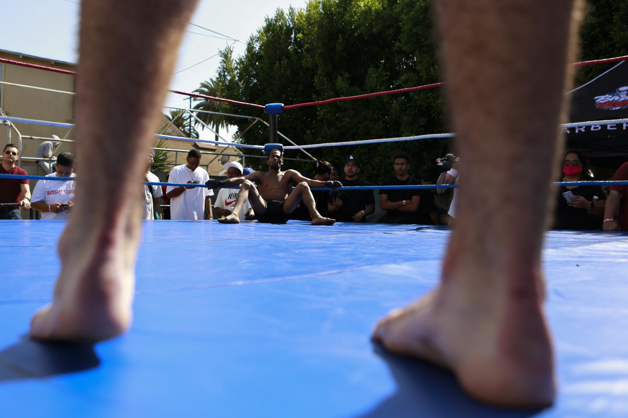 A man sits in a boxing ring, viewed through the bare legs of his opponent.