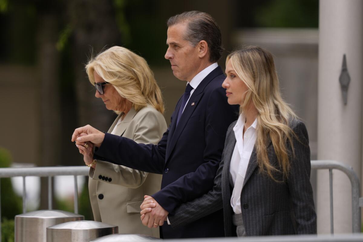 A dark-haired man in a dark suit and tie walks while holding hands with two women with blond hair flanking him