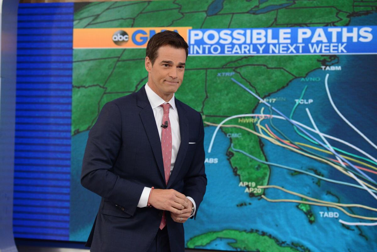 A weatherman in a dark suit and red tie stands with hands clasped in front of a map showing possible paths of a storm