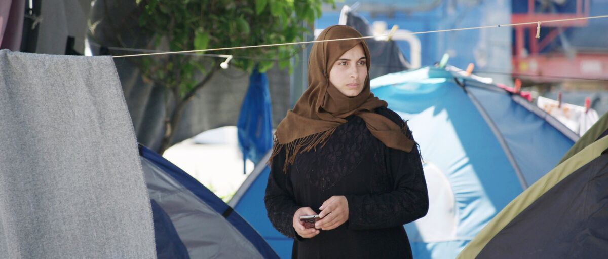 A woman in a headscarf stands among tents holding a phone 