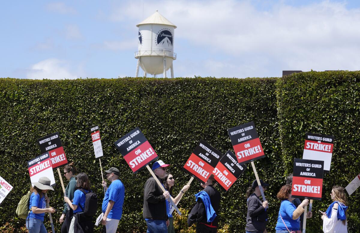 Striking writers march and hold signs in front of a large green hedge and a Paramount sign