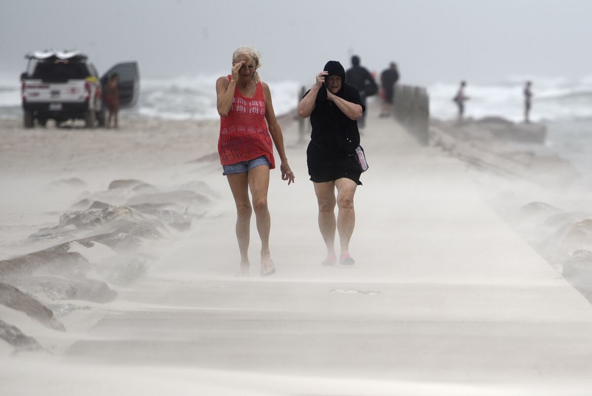 Two women walk beside a beach in a haze of sand. One holds a hand over her eyes; the other pulls up her hood.