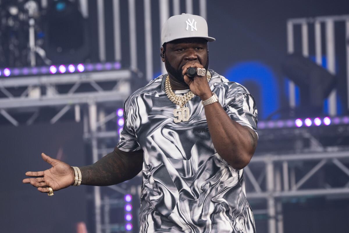 50 Cent in a gray hat, patterned shirt and gold chains holding a microphone to his mouth while performing onstage