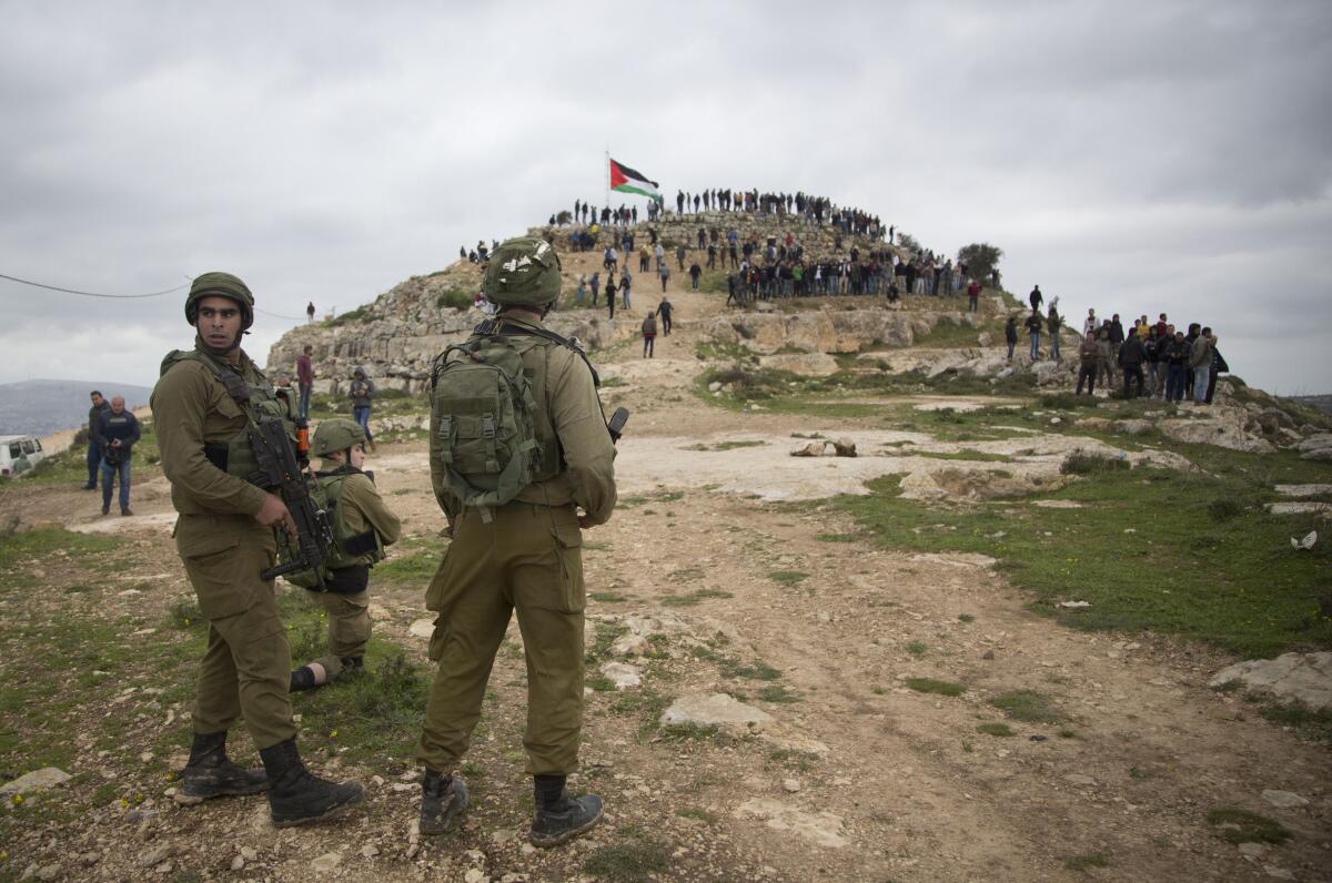 Israeli soldiers, foreground, monitor a crowd on a hilltop bearing a Palestinian flag 