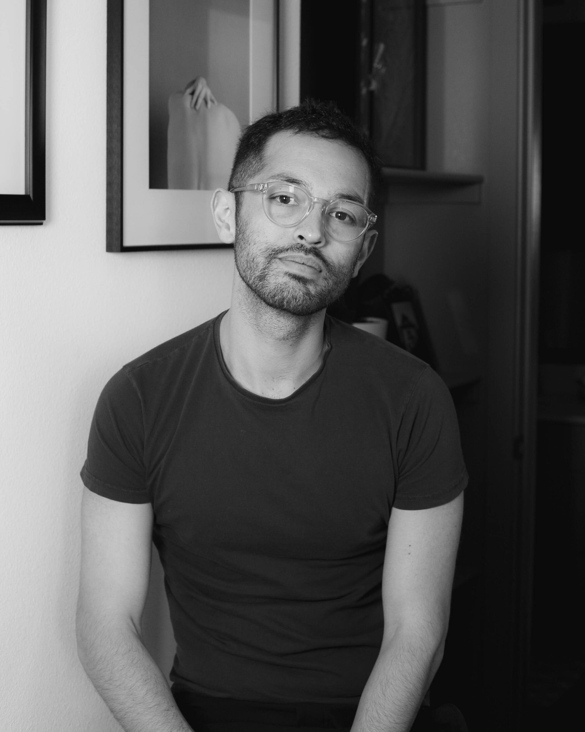 A man with clear glasses sits near a white wall with frames