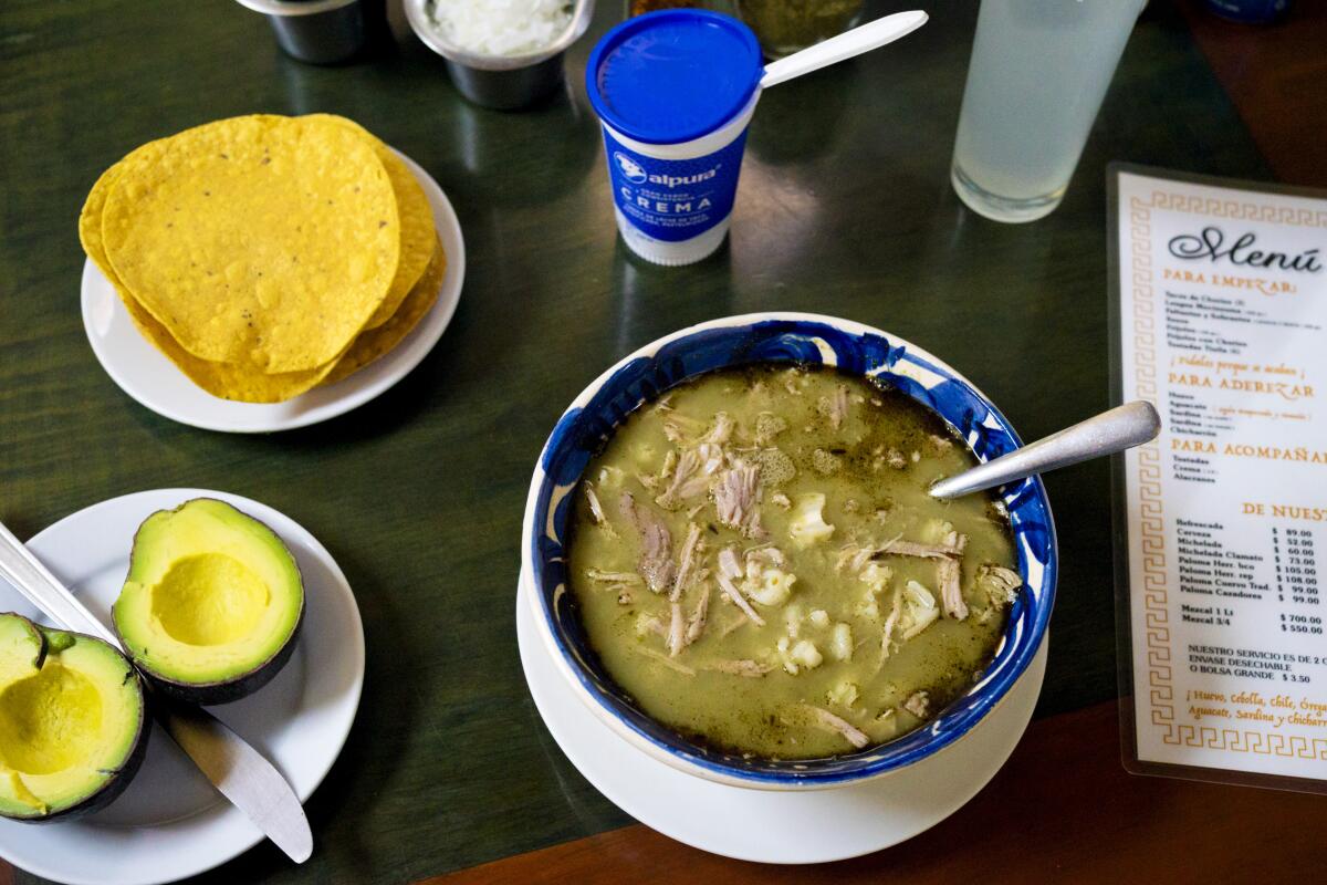 A dish of green-hued pozole next to an open avocado and tortillas.