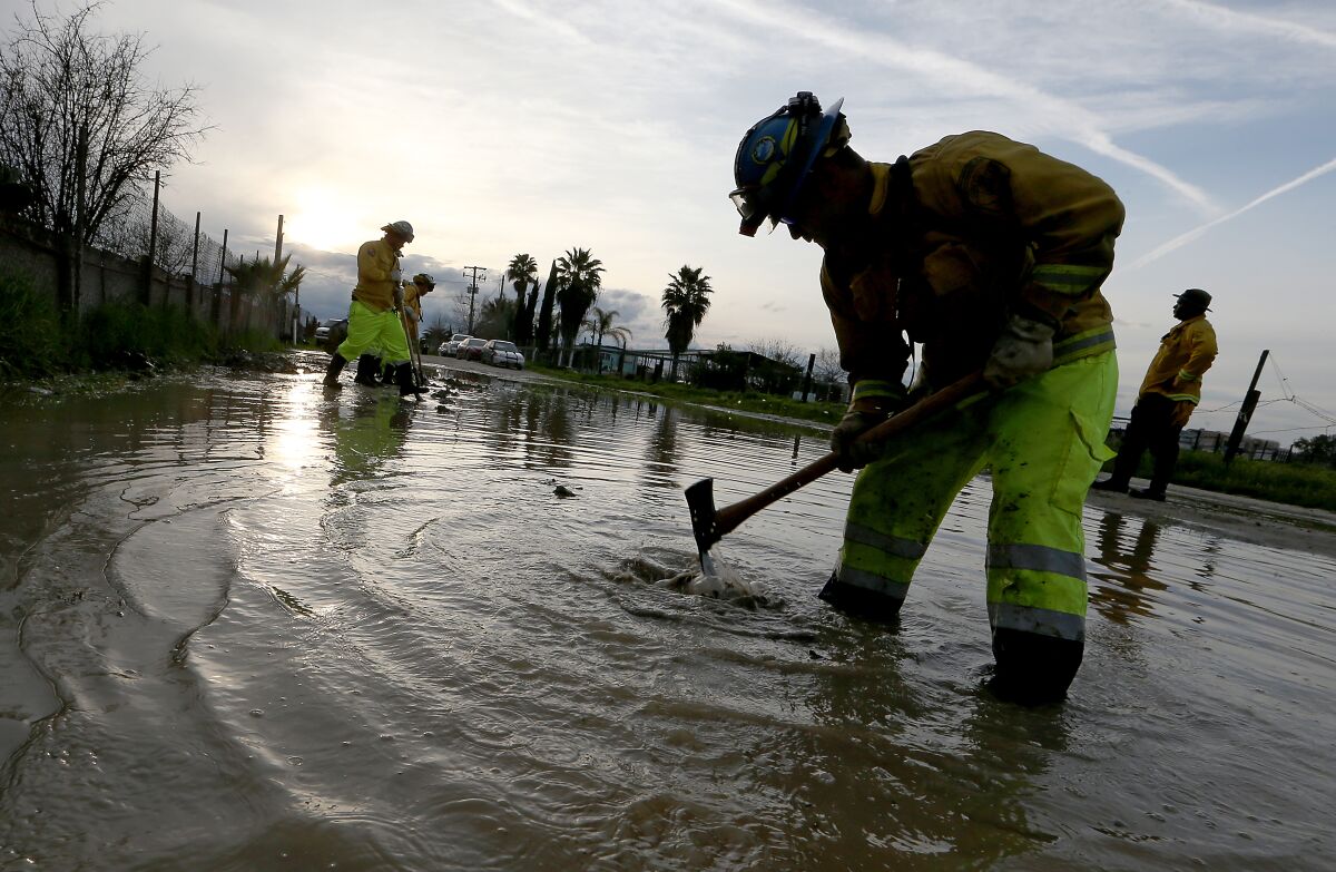 A firefighter uses a hand tool to dig into a flooded road with other crew members standing behind him.