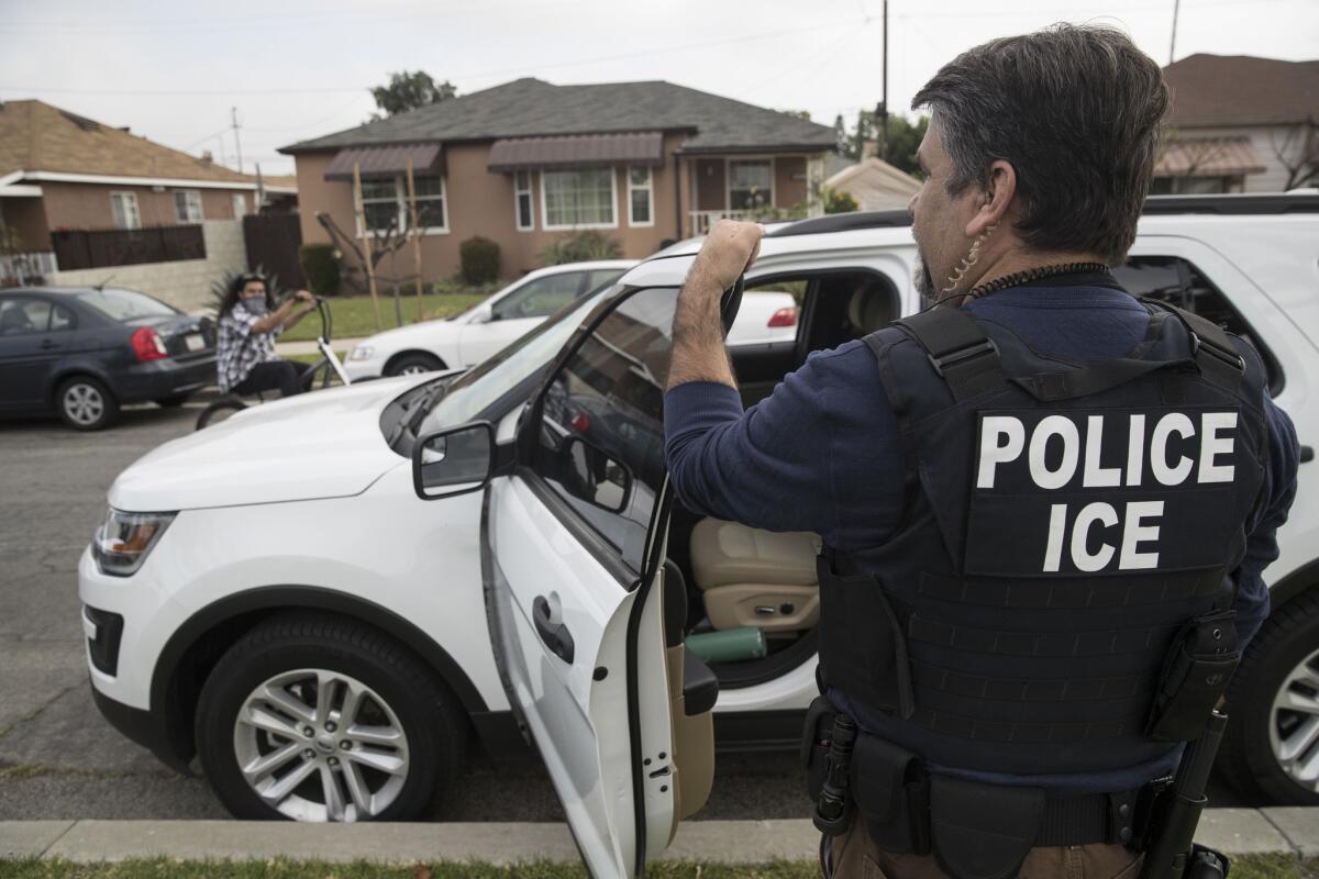 A man with a dark vest that says Police Ice, seen from behind, stands on a sidewalk holding the open door of a vehicle 