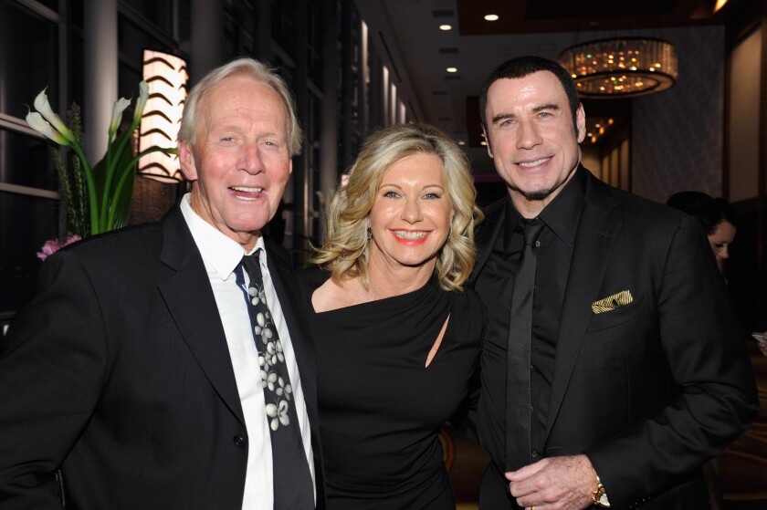 A smiling blonde woman in a black dress stands between two men in suits at a gala event