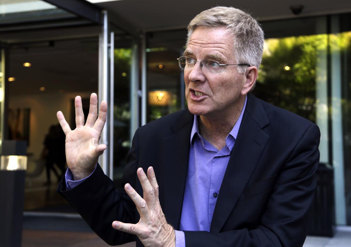 Rick Steves, wearing a blue shirt and dark blazer, speaks while motioning with his hands